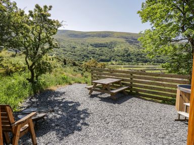 An outdoor patio with a wooden picnic table chairs fire pit and hot tub overlooking green hills at Moelfre - Shepherds Hut in Llanbedr