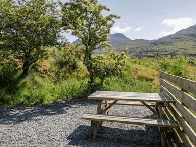 An outdoor picnic table on a gravel area near trees and greenery with mountains in the background at Moelfre - Shepherds Hut in Llanbedr