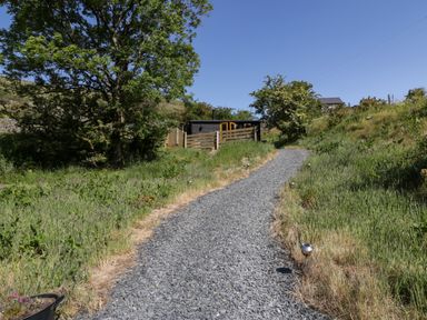 A gravel path leading to a wooden fenced shelter surrounded by grass and trees at Moelfre Shepherds Hut in Llanbedr