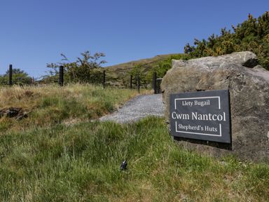 A gravel path leading through grass and bushes with a stone sign for Cwm Nantcol Shepherds Huts in Llanbedr