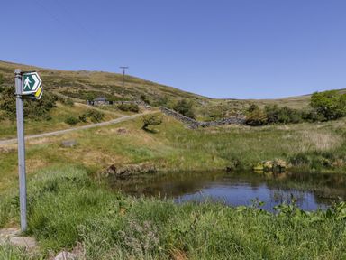A small pond with grass around it and a walking path with a walking signpost at Moelfre - Shepherds Hut in Llanbedr