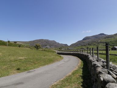 A country road with a stone wall and fence beside green fields and hills in the background at Moelfre - Shepherds Hut in Llanbedr