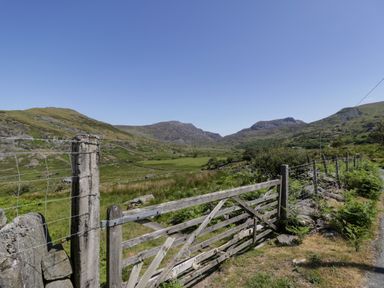 A wooden gate and wire fence along a rural road with hills and grassy fields in the background at Moelfre - Shepherds Hut in Llanbedr