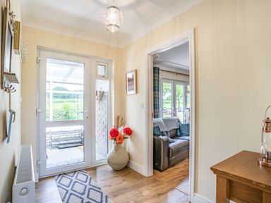 An entryway with a glass door leading outside a large vase with flowers on the floor and a view into a living room with a leather sofa at Yr Hen Ffald in Pontarddulais