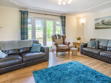 A living room with two leather sofas an armchair a blue rug wooden tables and French doors with blue curtains at Yr Hen Ffald in Pontarddulais