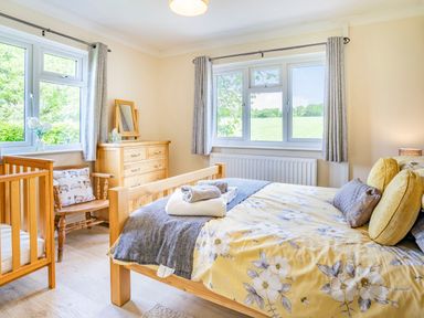 A bedroom with a wooden bed with floral bedding and towels on top a wooden crib a wooden chair with a sheep pillow a wooden dresser with a mirror and two windows with curtains at Yr Hen Ffald in Pontarddulais