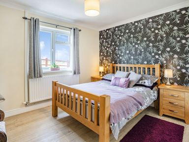 A bedroom with a wooden double bed with grey and purple bedding two wooden nightstands with lamps a window with grey curtains and floral wallpaper at Yr Hen Ffald in Pontarddulais