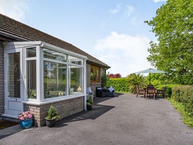 An outdoor patio area with a glass entrance porch potted plants outdoor chairs a table with an umbrella and surrounding greenery at Yr Hen Ffald in Pontarddulais