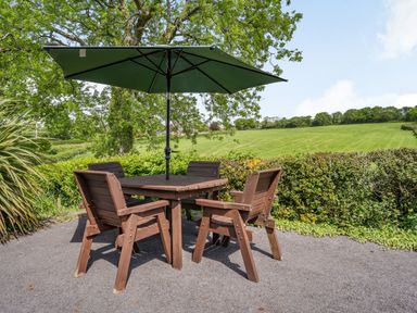 A wooden outdoor table with four chairs and a green umbrella on a paved area overlooking a grassy field at Yr Hen Ffald in Pontarddulais