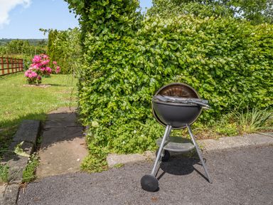 A barbecue grill on wheels on a driveway next to a hedge and a garden with flowers and grass at Yr Hen Ffald in Pontarddulais