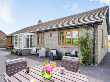 An outdoor seating area with wooden table and chairs a vase of flowers and two cups in front of a gray brick house with windows and patio furniture at Yr Hen Ffald in Pontarddulais