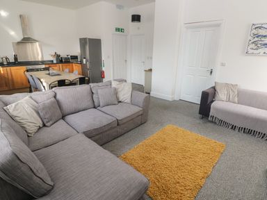 A living room with grey sofas, a yellow rug, and an open kitchen with wooden cabinets and a dining table at The Shell at the Hideaway in Tenby