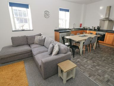 A living area with a grey sofa and a small wooden side table next to a dining table with chairs in a kitchen at The Shell at the Hideaway in Tenby
