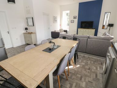 A dining area with a light wooden table and chairs next to a living room with gray sofas and a television on a cabinet at The Shell at the Hideaway in Tenby