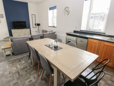A dining area with a wooden table and chairs next to a kitchen with appliances and a living room with a gray sofa and television at The Shell at the Hideaway in Tenby