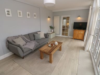A living room with a grey sofa pillows and a blanket a wooden coffee table with tea set a wooden cabinet with a television and framed pictures on the wall at Castle at The Hideaway in Tenby
