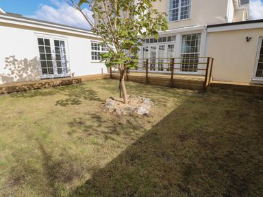 A backyard with a small tree surrounded by rocks and grass and a white house with multiple glass doors and windows and a wooden deck at Castle at The Hideaway in Tenby