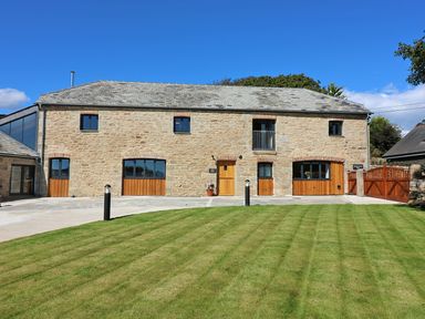 A stone building with wooden doors and windows a grass lawn and paved driveway at Mill House Boskensoe Barns in Mawnan Smith near Falmouth