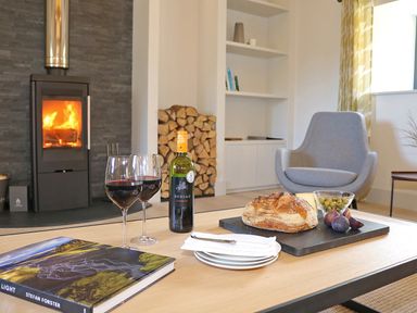 A living room with a wood burning stove and logs near a coffee table with wine glasses bread and fruit at Mill House Boskensoe Barns in Mawnan Smith near Falmouth