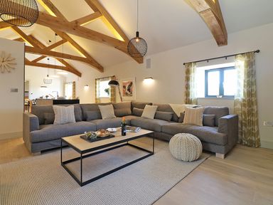 A living room with a large gray sectional sofa a wooden coffee table and exposed wooden ceiling beams at Mill House Boskensoe Barns in Mawnan Smith near Falmouth