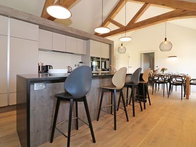 A kitchen with a breakfast bar with stools and a dining table in the background at Mill House Boskensoe Barns in Mawnan Smith near Falmouth
