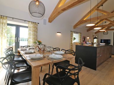 A dining area with a wooden table set with plates and glasses and black chairs near kitchen island with pendant lights at Mill House Boskensoe Barns Mawnan Smith near Falmouth