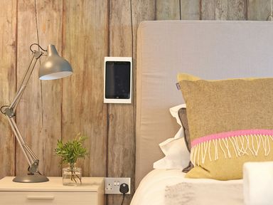 A bedside table with a lamp and a glass vase with greenery next to a bed with cushions and a wooden wall behind at Mill House Boskensoe Barns in Mawnan Smith near Falmouth