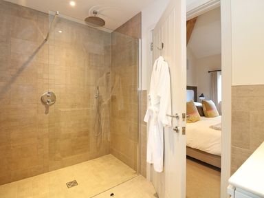 A bathroom with a walk-in shower and a white bathrobe hanging on the door showing a bedroom with a bed and pillows at Mill House Boskensoe Barns in Mawnan Smith near Falmouth