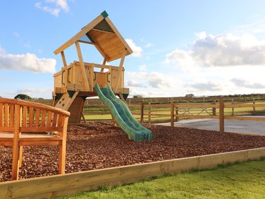 A wooden children's play structure with a green slide and a wooden bench in an outdoor fenced area Mill House Boskensoe Barns Mawnan Smith near Falmouth