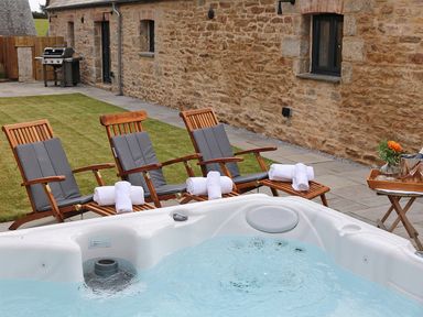 An outdoor area with a hot tub wooden lounge chairs with gray cushions and towels a tray with flowers and drinks a stone wall and a barbecue grill at Mill House Boskensoe Barns Mawnan Smith near Falmouth