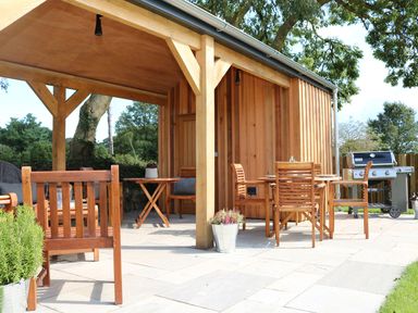An outdoor patio area with wooden chairs and tables under a wooden canopy near a barbecue grill at Mill House Boskensoe Barns Mawnan Smith near Falmouth
