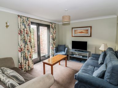 A living room with two sofas a blue armchair a wooden coffee table a flat screen TV on a stand floral curtains and a framed picture at The Lodge off High Street in Hadleigh