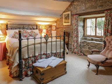 A bedroom with a metal bed frame wooden chest armchair stone walls and floral curtains at Lower Dolgenau- The Cottage in Llawryglyn near Llanidloes