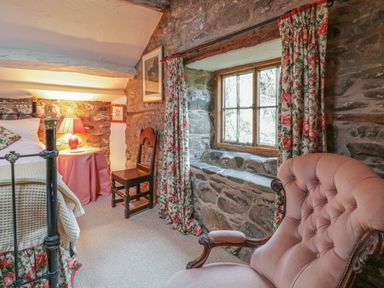 A bedroom with a pink armchair floral curtains stone walls a wooden window a wooden chair and a bedside table with a lamp at Lower Dolgenau- The Cottage in Llawryglyn near Llanidloes