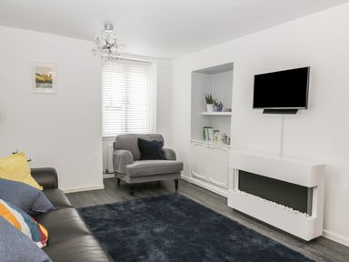 A living room with a gray armchair, a dark sofa with cushions, a wall-mounted TV, and a modern white fireplace at 11 Camden Building in Kendal