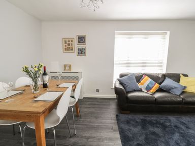 A dining table with chairs and a bottle of wine next to a sofa with cushions and a window at 11 Camden Building in Kendal