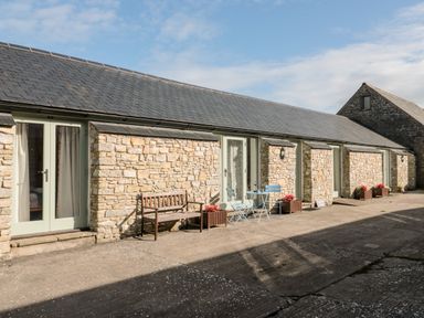 A stone building with green doors a wooden bench a blue table with chairs and flower pots outside at Poppy Cottage in Llantwit Major