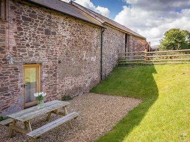 A stone building with a wooden door a picnic table with a vase of flowers on gravel next to a grassy area at Salters in Black Dog near Crediton