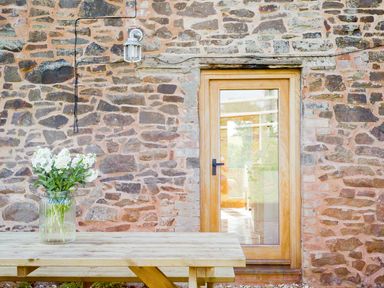 A wooden outdoor table with a glass jar of white flowers against a stone wall with a wooden door and a wall light at Salters in Black Dog near Crediton