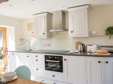 A kitchen with white cabinets, a black countertop, a built-in oven, a stove, a sink with a drying rack, a kettle, a toaster, an egg cup with an egg, a loaf of bread on a cutting board, two cups on saucers, a teapot, a cutting board, a plant, and a fire extinguisher near a wooden door at Salters in Black Dog near Crediton