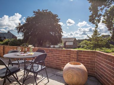 A patio with a round metal table and four chairs with a bottle and glasses on the table and a wooden planter against a brick wall at Salters in Black Dog near Crediton