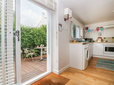 A kitchen with white cabinets and a washing machine near a sliding glass door opening to a wooden deck with two metal chairs and a round table at Bolitho's Retreat in Helford