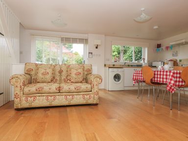 An open plan room with a floral sofa wooden floor dining table with red and white checkered tablecloth chairs kitchen appliances and windows showing greenery outside at Bolitho's Retreat in Helford