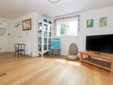 A room with a wooden floor a television on a wooden table a white display cabinet a blue chair a small black table and a window with floral curtains at Bolitho's Retreat in Helford