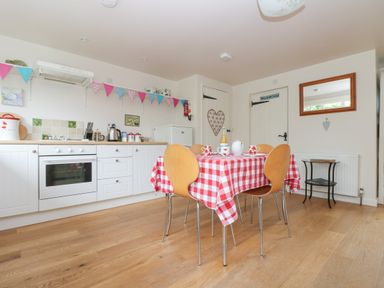 A kitchen with white cabinets and an oven with a red and white checkered tablecloth on the table and wooden chairs at Bolitho's Retreat in Helford