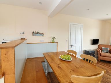 A dining area with a wooden table and chairs a bowl of green apples a built in wooden bench a beige armchair a small black stove and a wall mounted television at Bolenowe in Trebetherick near Polzeath