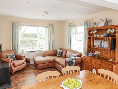A living room with brown leather armchair and sectional sofa next to wooden cabinet with dishes and a dining table with a bowl of apples at Bolenowe in Trebetherick near Polzeath