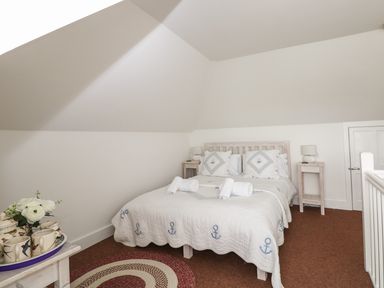 A bedroom with a double bed covered by a white quilt with blue anchors and matching pillows two wooden bedside tables with lamps a small round rug and a small table with cups and flowers at Bolenowe Trebetherick near Polzeath