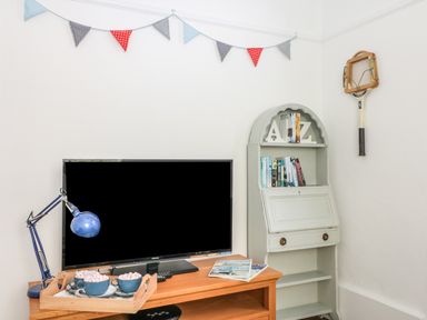 A television on a wooden cabinet with a blue lamp and a tray with mugs beside a white bookshelf and a vintage tennis racket on the wall at Bolenowe Trebetherick near Polzeath