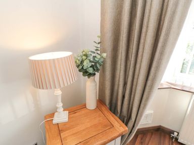 A wooden table with a white lamp and a vase with green foliage next to a beige curtain at Lilly May Cottage in Flamborough